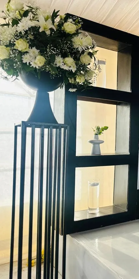 A white floral arrangement in a black urn on a slatted pedestal beside glass candle holders